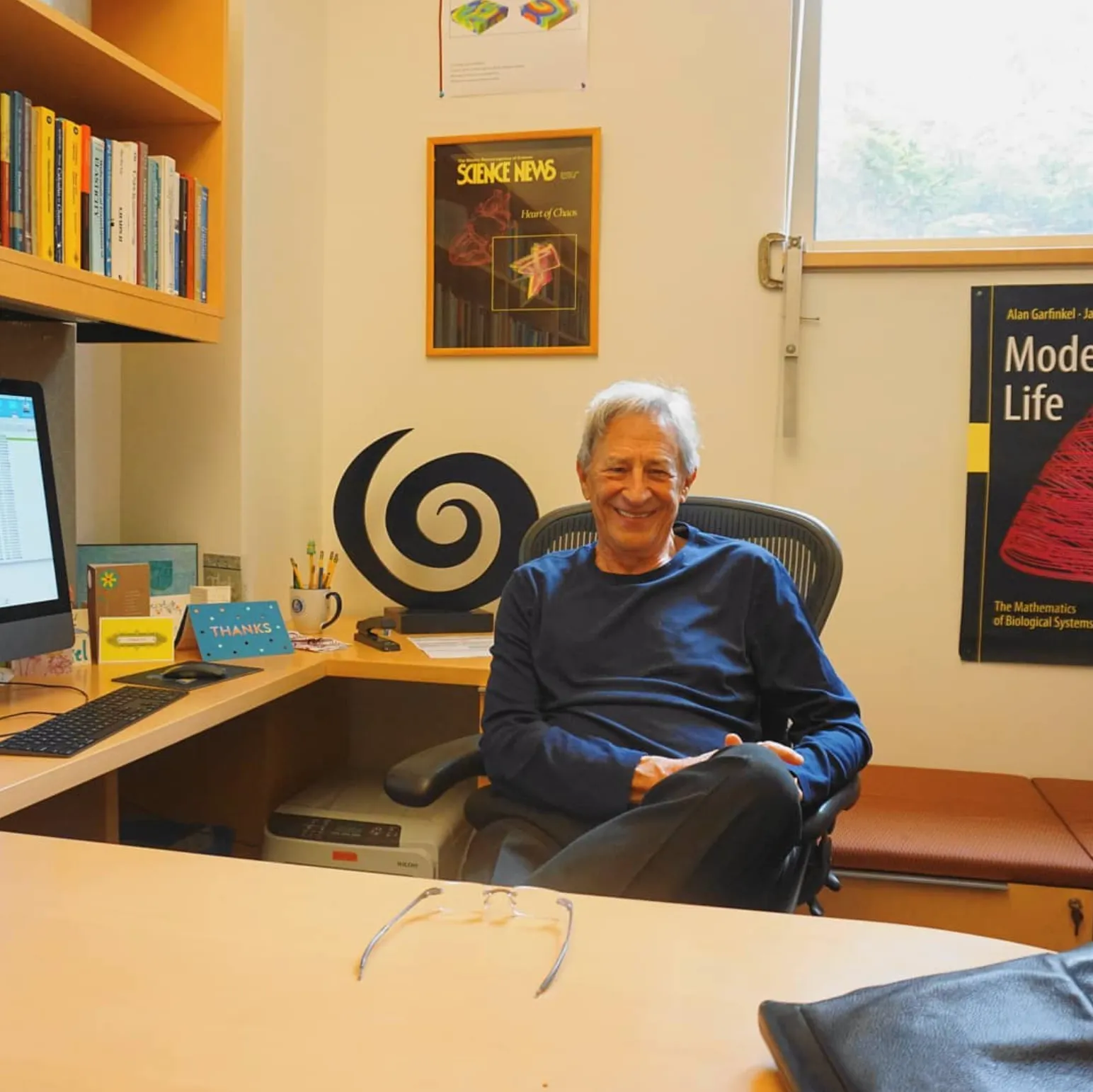 Alan Garfinkel smiling in his office, seated at his desk with bookshelves, a Science News cover on chaos theory, and his book Modeling Life on the wall